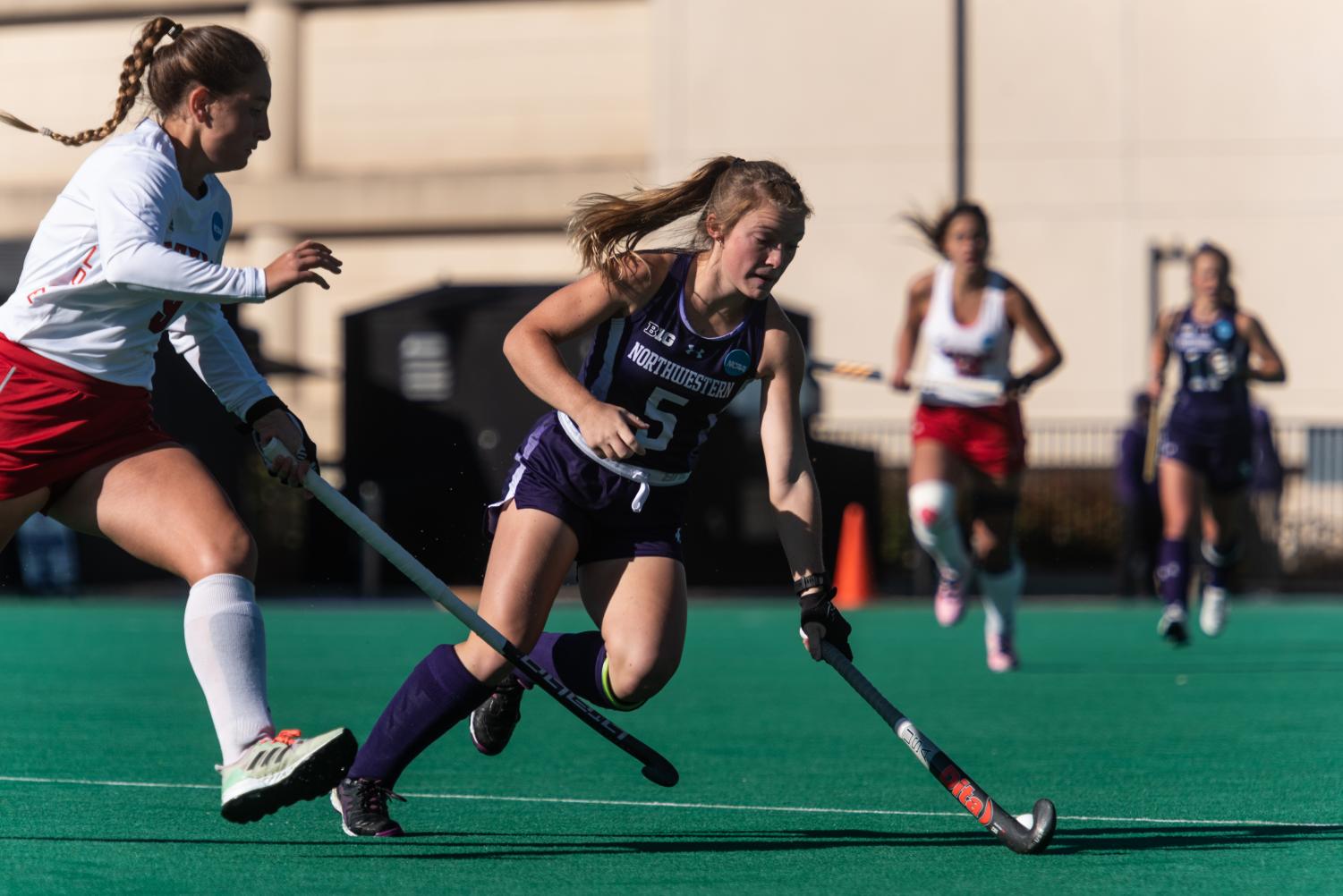 A field hockey player in a purple jersey with the number five runs ahead and uses the stick to keep the ball away from a player in a white jersey.