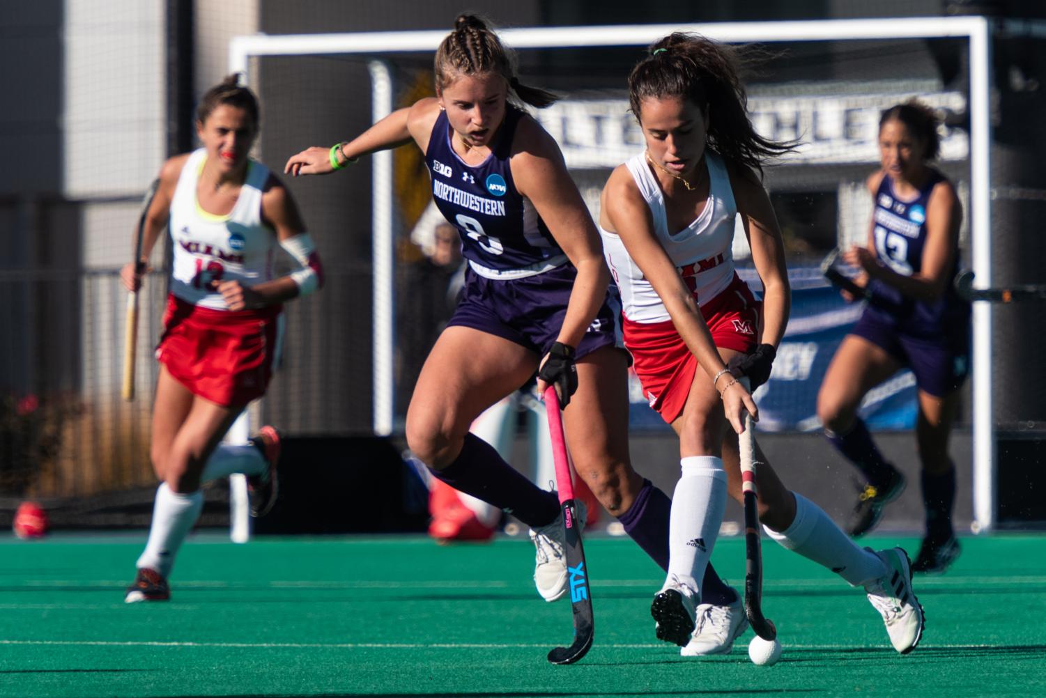 A field hockey player in a purple jersey chases alongside a player in a white jersey who has the ball under their stick.