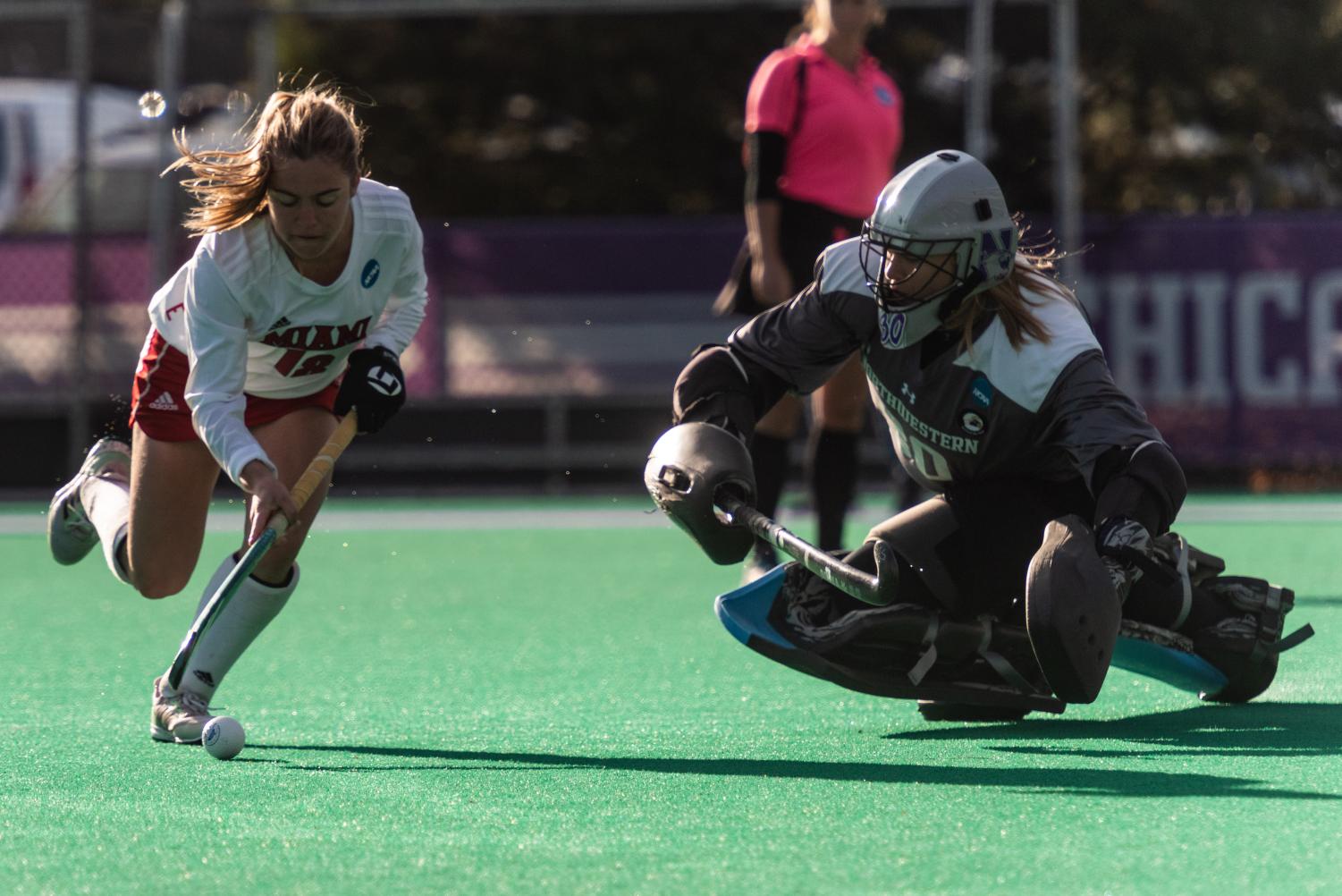 A field hockey goalie is down on their knees to defend a goal being scored from a ball being hit by a player in a white jersey.