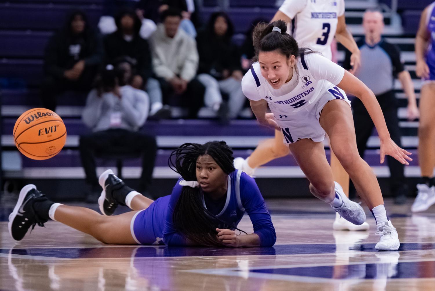 A basketball player in purple is flat on the ground and another player to their right in white is running towards them. Both players look at the basketball to their right.