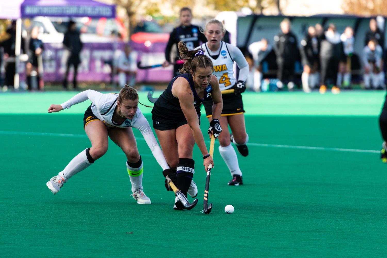 An athlete in a black jersey chases after a white ball with a field hockey stick as an athlete in white jersey uses a field hockey stick to reach for the ball.