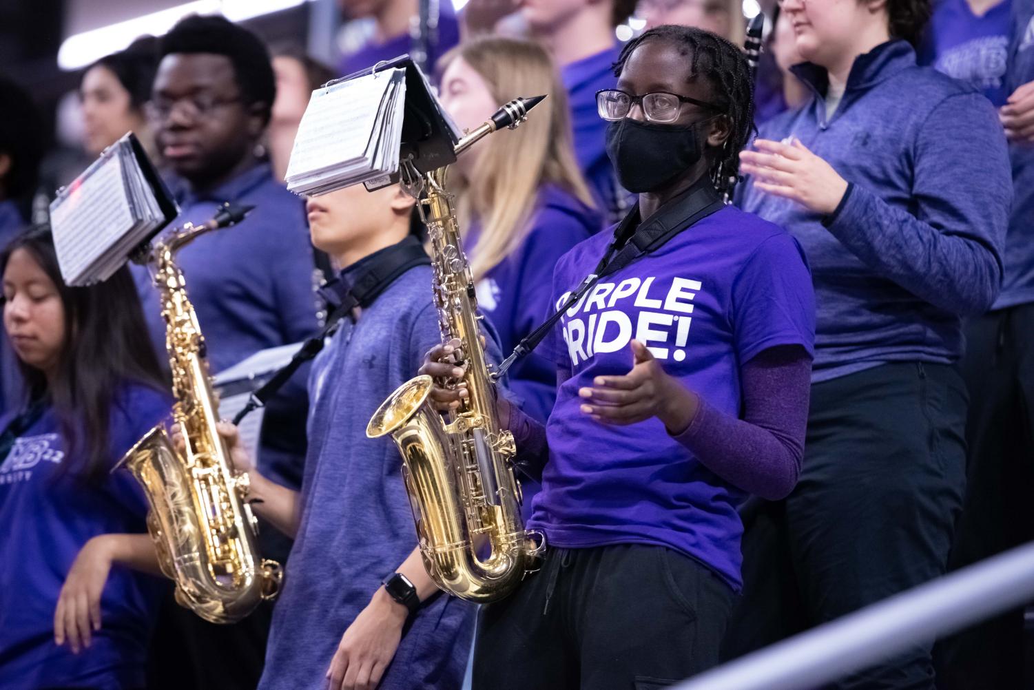 Band players in purple play brass instruments. The person in the foreground with a black mask holds a saxophone.