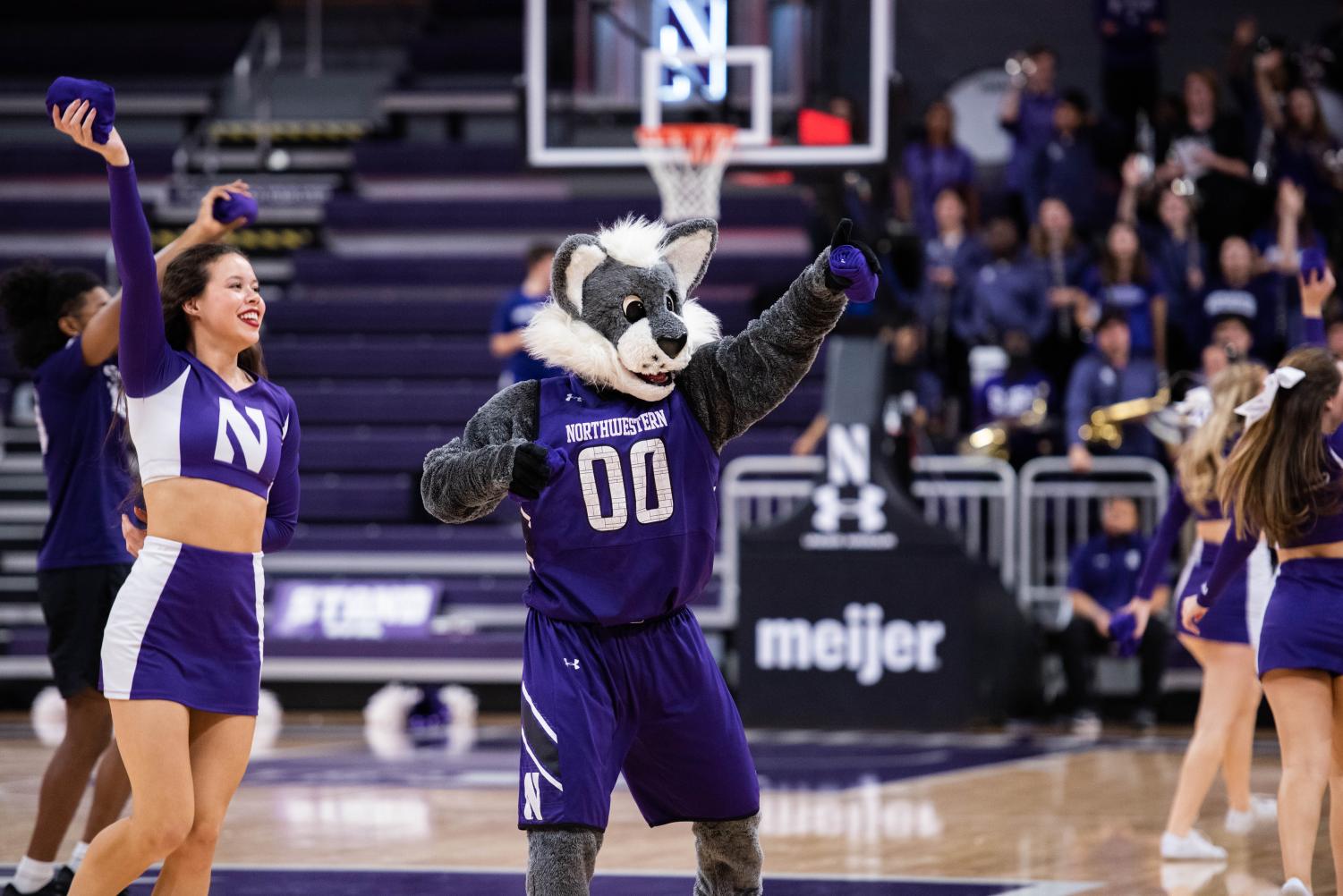 A person in a wildcat costume and a purple jersey holds a rolled-up t-shirt and points.