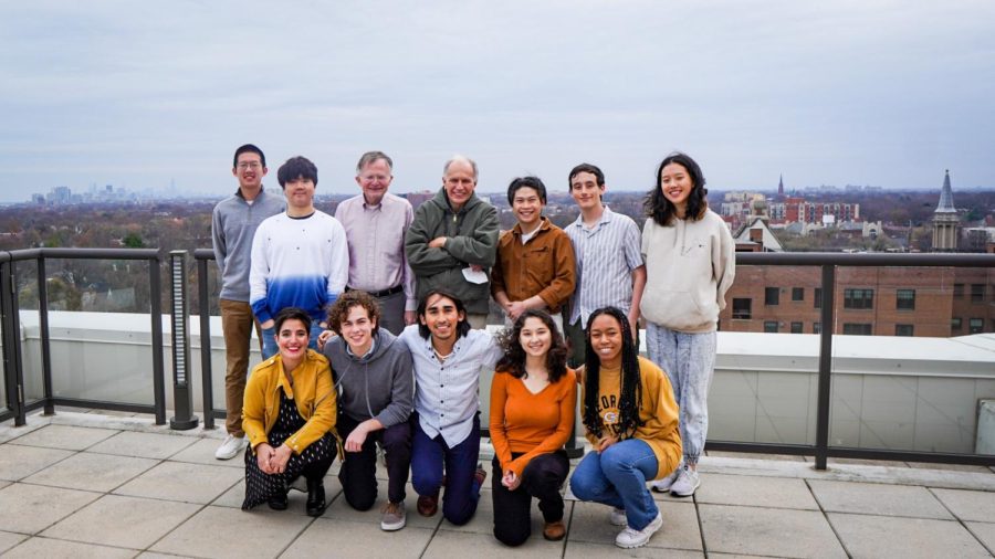Students and other Thanksgiving dinner attendees stand on a rooftop patio overlooking Evanston, with Chicago in the background in the left of the picture. There are 12 attendees standing in two lines, with the back row standing and the front row crouching.