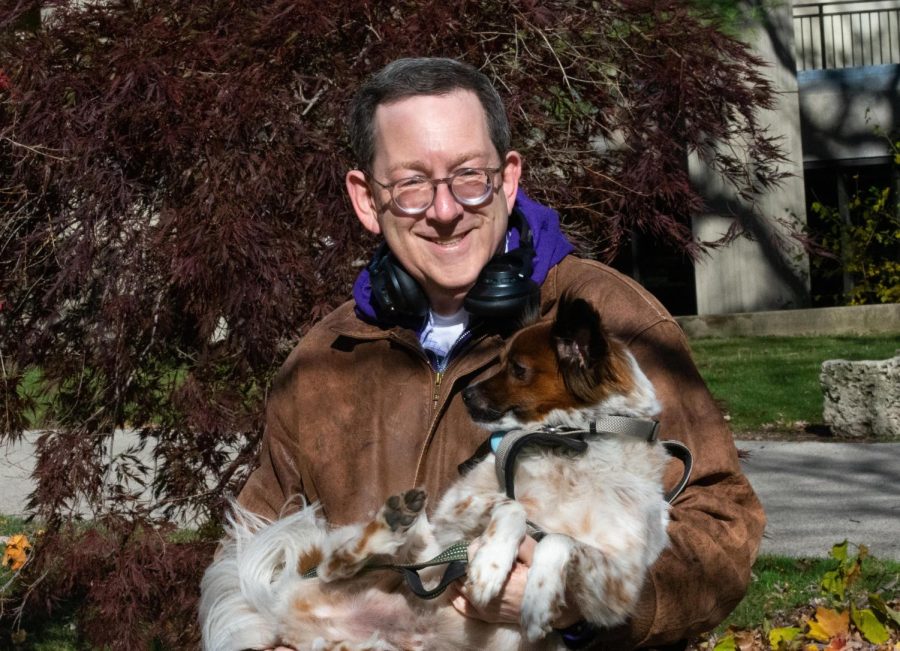 A person wearing a dark brown jacket with a purple hoodie holds his brown and white dog