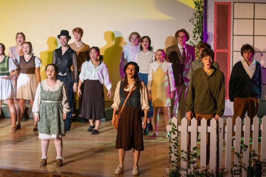 An ensemble of people stand onstage and sing behind a white picket fence set piece.
