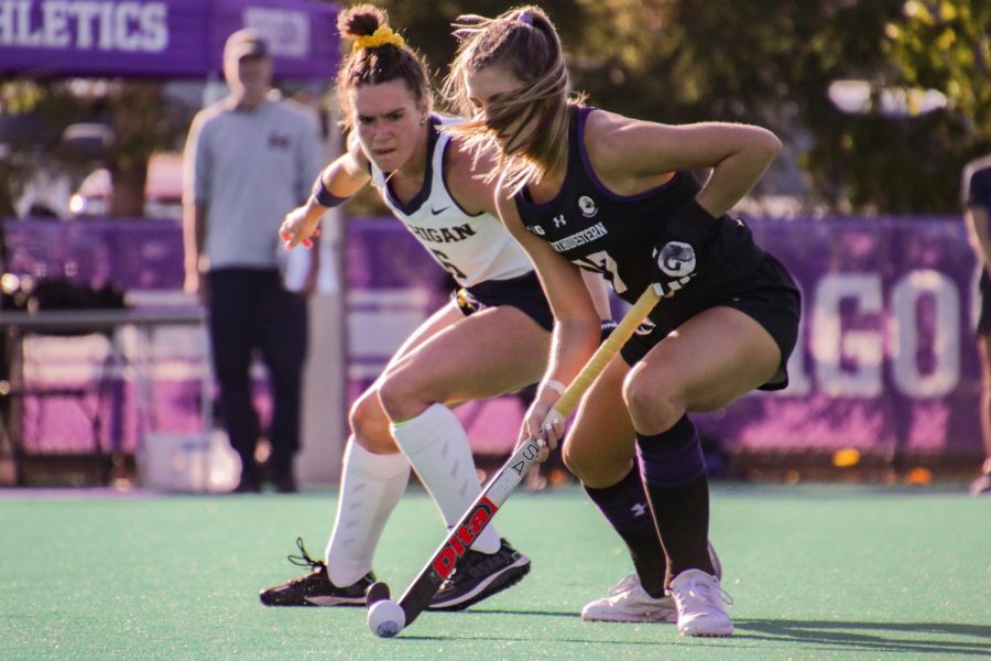 An athlete in a black jersey pushes a ball with a field hockey stick while an athlete in a white jersey looks on.