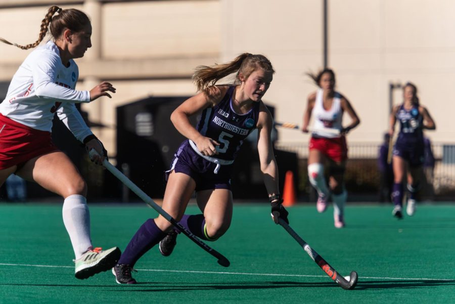An athlete in a purple jersey hits a ball with a field hockey stick.