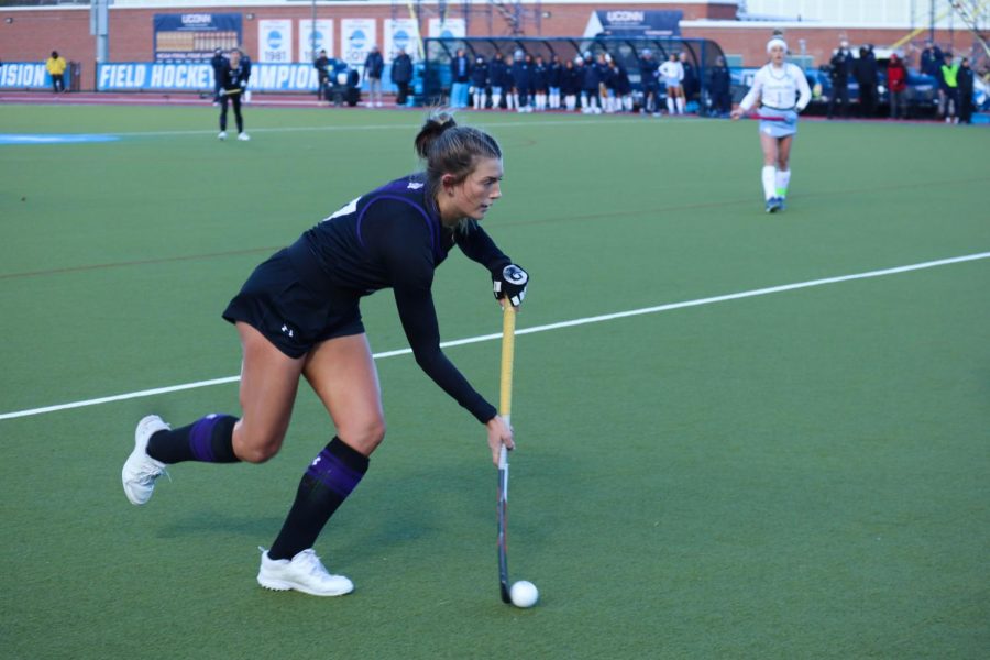 Field hockey player in black uniform runs down field with the ball on her stick.