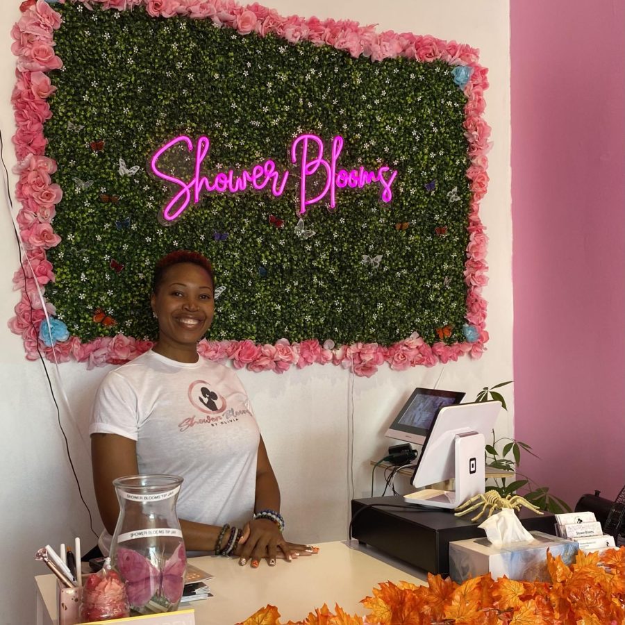 Person smiling at the camera as she stands before a green vine wall bordered with large pink flowers and a glowing neon pink sign in the middle that reads “Shower Blooms.”