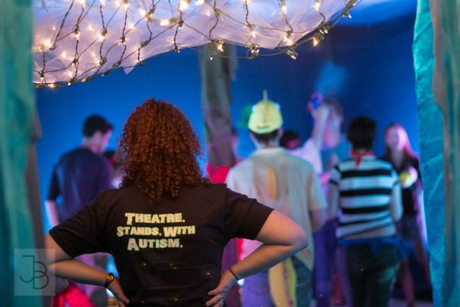 A person wearing a “Theatre Stands with Autism” T-shirt faces away from the camera. The set of a theatre performance is in the background.