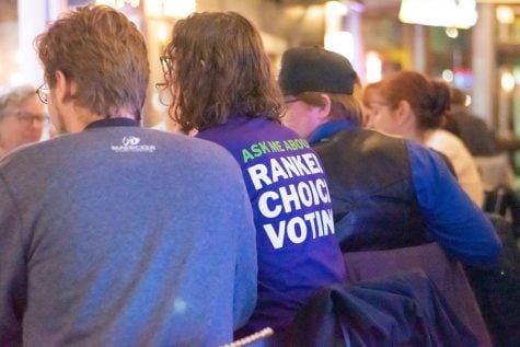 Members of RCV for Evanston sit at a table pictured from behind. One person’s shirt reads: “ASK ME ABOUT RANKED CHOICE VOTING”