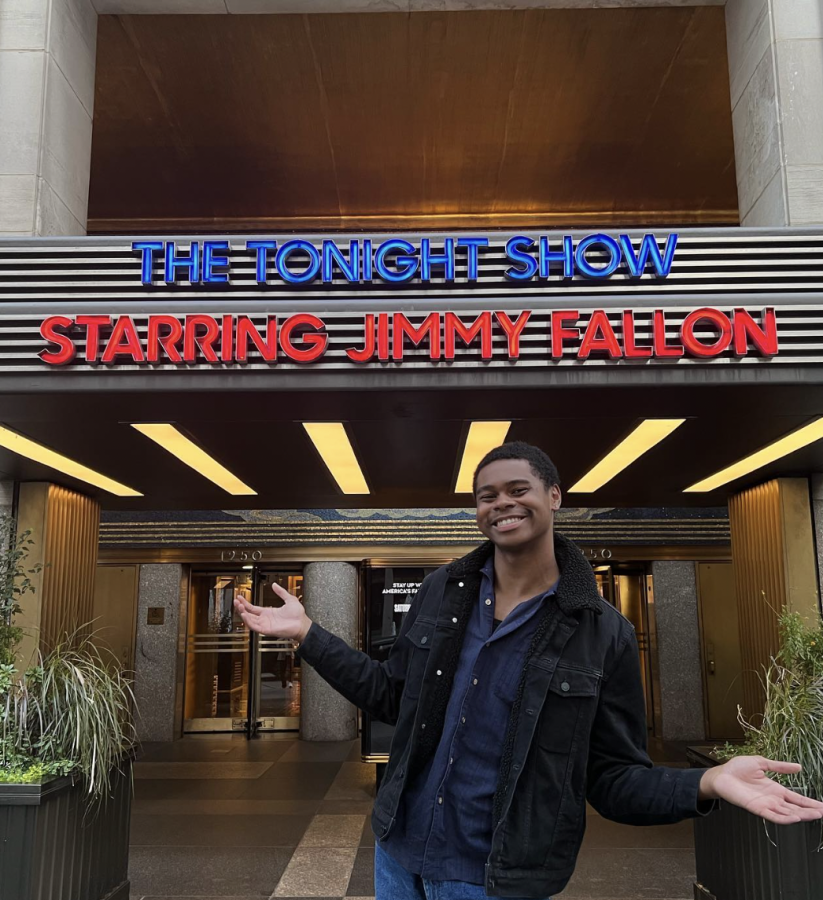 A man wearing a black jacket and blue pants stands in front of a building with a sign that reads, “The Tonight Show/ Starring Jimmy Fallon.”