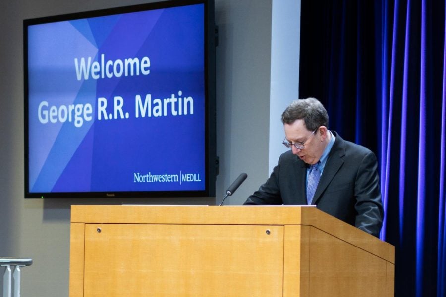 University President Michael Schill stands at a podium in front of purple curtains, and a screen reads, “Welcome George R.R. Martin.”