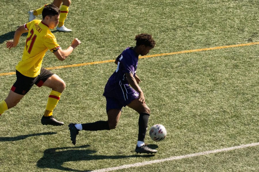 A soccer player in a purple jersey and purple shorts dribbles a ball.
