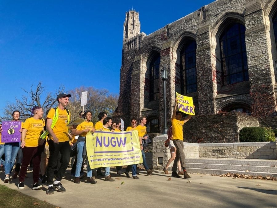 People walk carrying yellow banners and posters in front of a stone building with a blue sky in the background.
