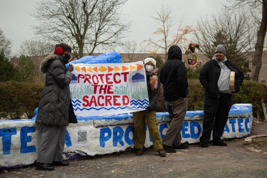 Two people holding up a handmade sign reading “Protect the Sacred” next to The Rock with a winter sky in the background.