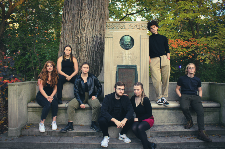 Seven+people+wearing+black+clothing+pose+in+front+of+a+concrete+Shakespeare+monument+with+trees+in+the+background.