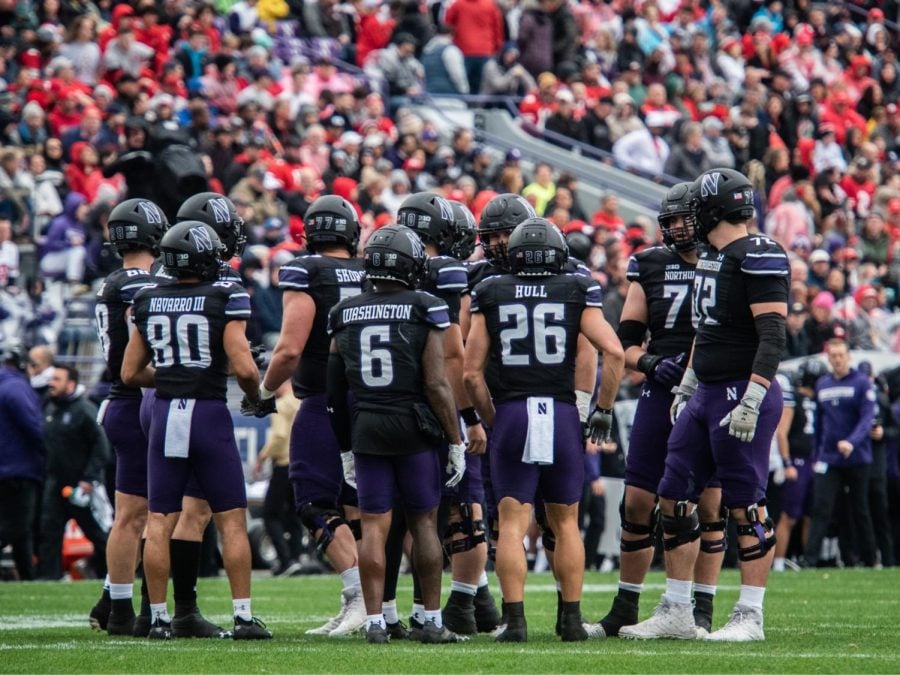 Football players in black jerseys, purple pants and black helmets huddle together.