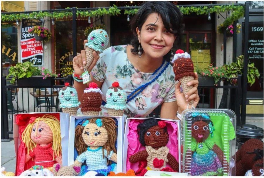 Woman+with+black+hair+in+a+floral+shirt+holds+up+two+crocheted+ice+cream+cones+and+poses+with+a+table+of+crocheted+dolls.