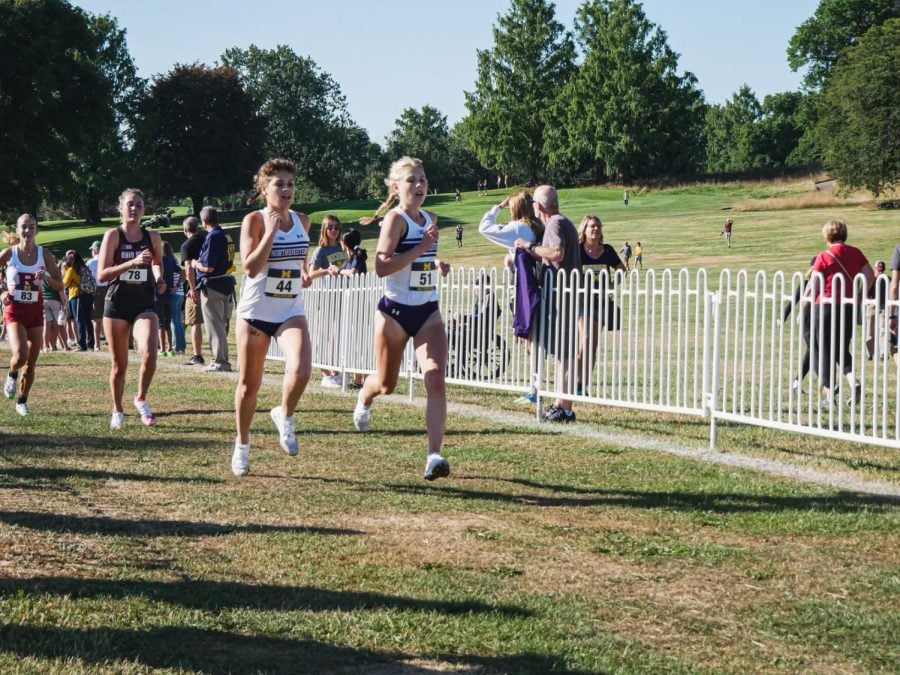 Two girls in white jerseys run next to each other in a cross country meet.