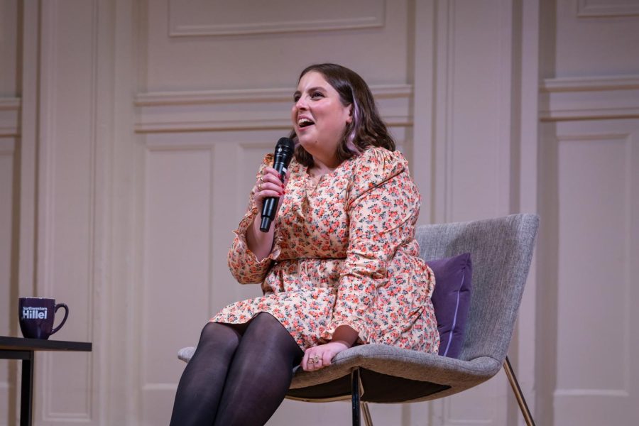 Beanie+Feldstein+sits+on+a+gray+chair+holding+a+microphone+and+wearing+a+floral+dress.
