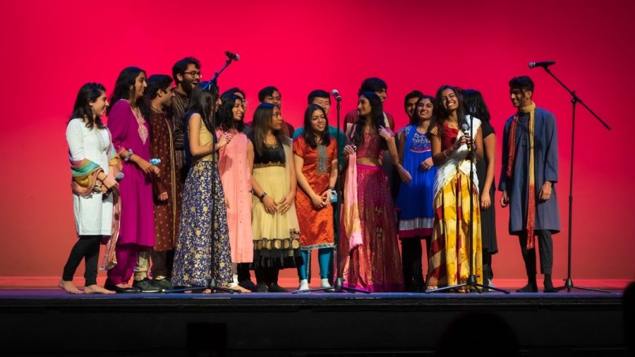 A+group+of+singers+on+a+stage+in+traditional+Indian+dress+gathered+around+microphones.
