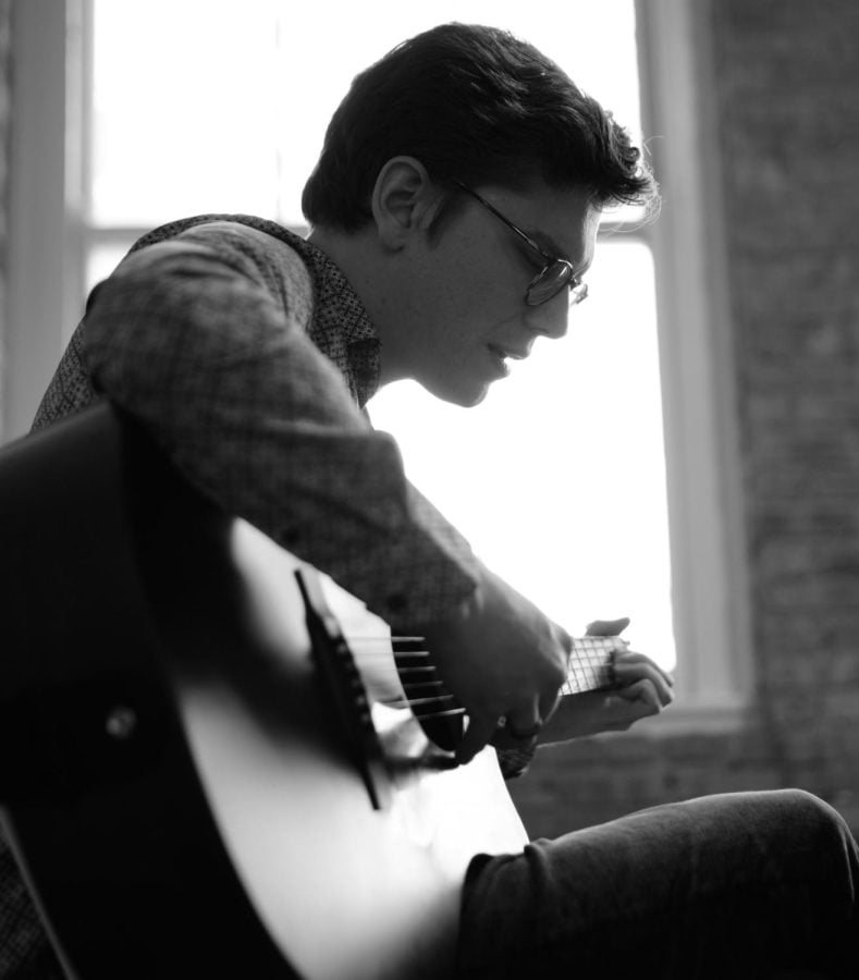 A man with short hair and glasses, wearing a checkered shirt plays guitar in front of a window.