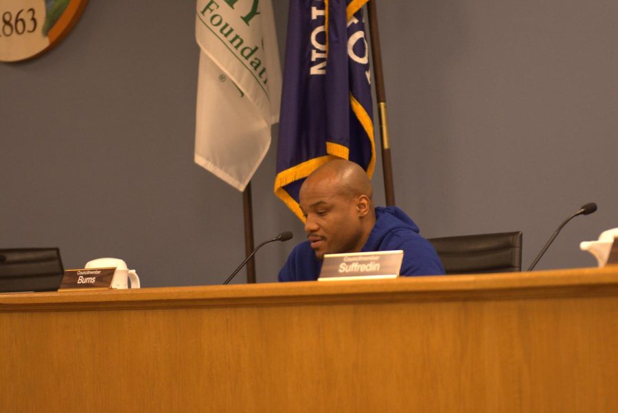 A councilmember wearing blue sits in front of flags at a wooden dais.