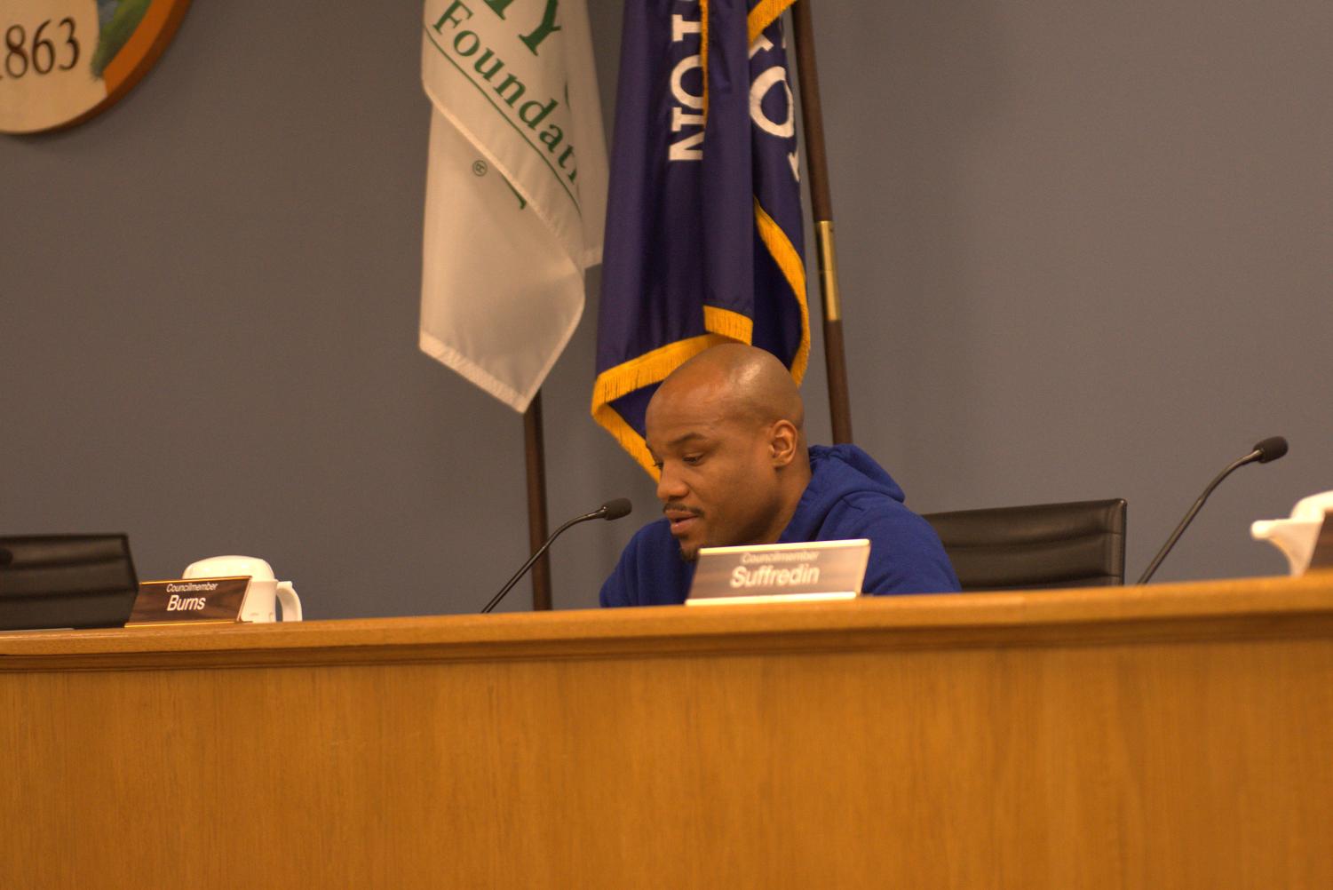 A+councilmember+wearing+blue+sits+in+front+of+flags+at+a+wooden+dais.