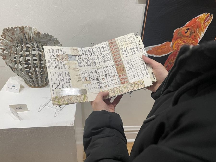 An attendee in a black jacket holds up a handmade, textured paper book. In the background, a shell-like vase is to the left and a black and orange weaving is seen to the right.