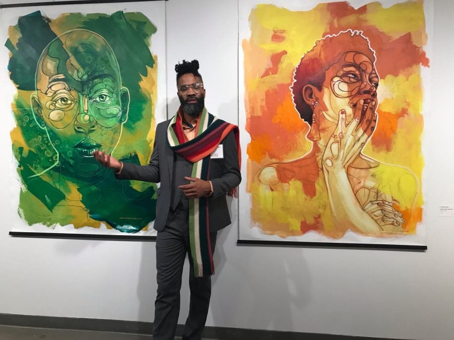 A person with a gray suit, a colorful scarf, a beard, and glasses stands in front of two large canvas portraits, a yellow and orange one on the right and a predominantly green painting on the left.