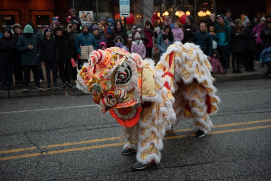 Two people in a lion costume dance in a street while a crowd watches.