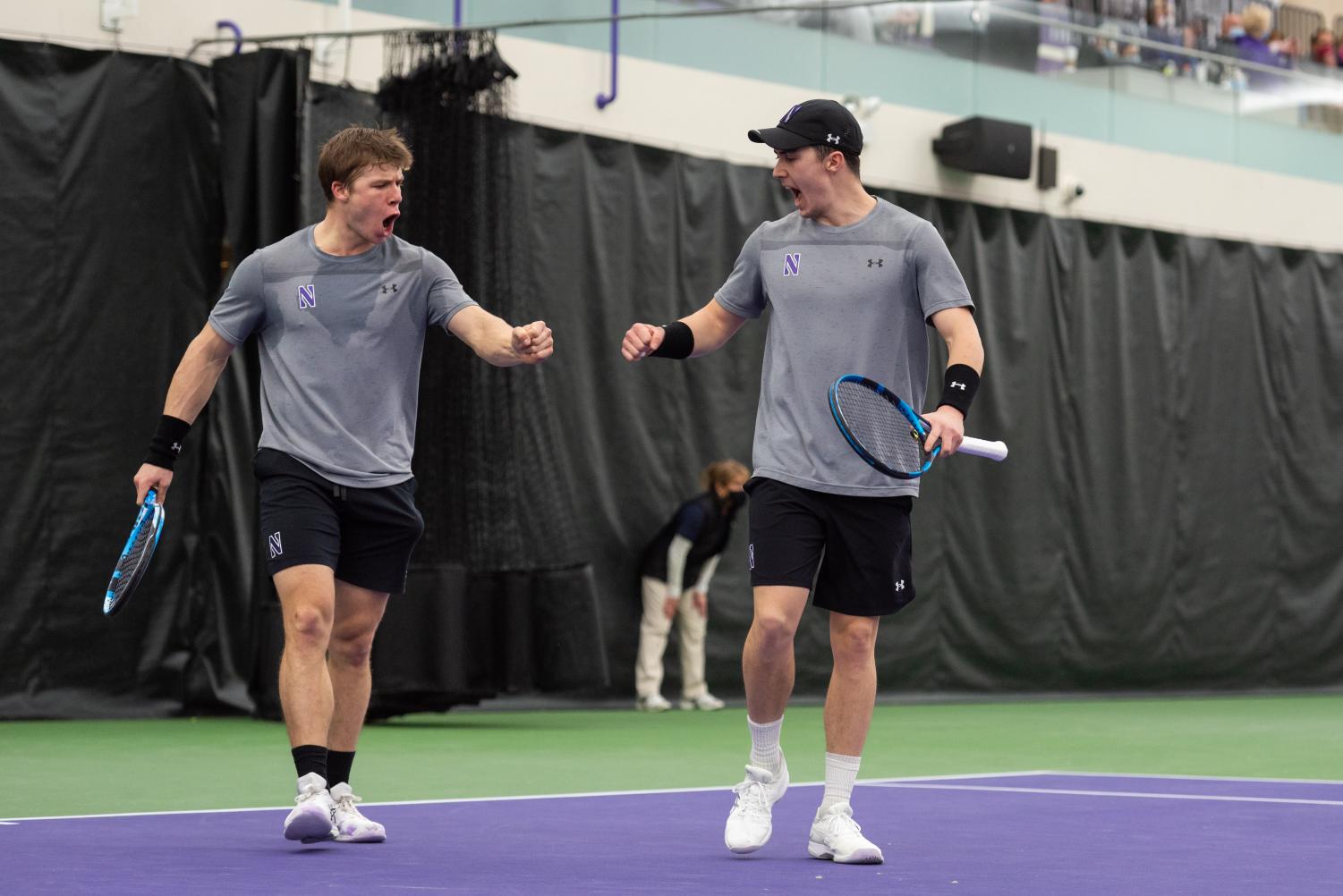 Two+tennis+players+in+gray+shirts+and+black+shorts%2C+each+holding+a+racquet%2C+fist+bump.