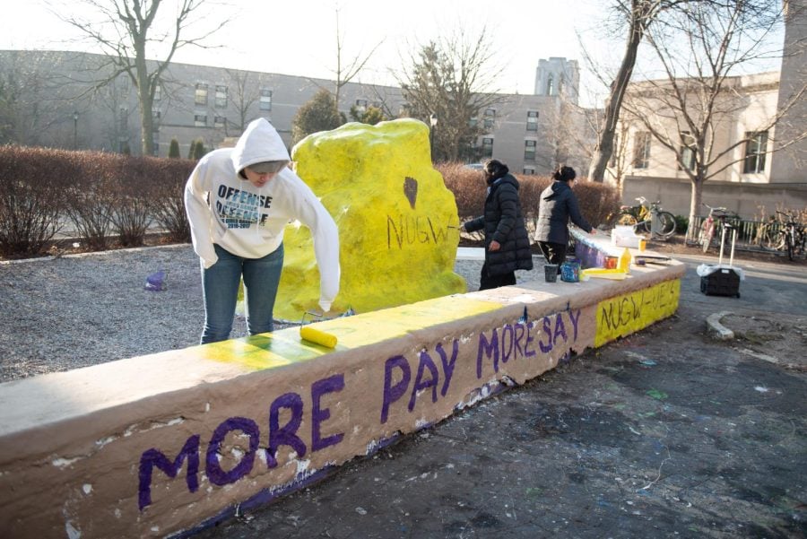 Three students paint a large yellow rock with the letters “NUGW.” A gray ledge in front of the rock is painted with “More Pay, More Say.”