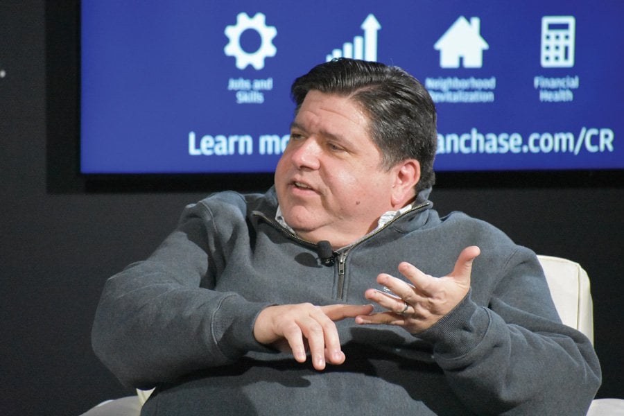 Gov. J.B. Pritzker, wearing a gray jacket, speaks at a meeting in front of a blue screen.