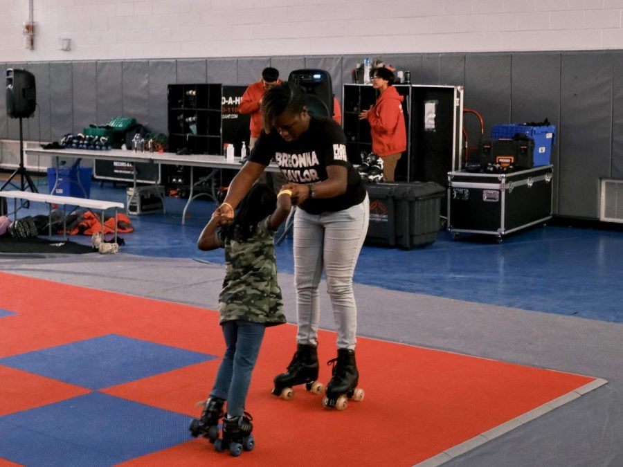A woman is holding the hands of a child, both standing on a red-and-blue rink and wearing roller skates. The woman is wearing a black T-shirt with the words Breonna Taylor.