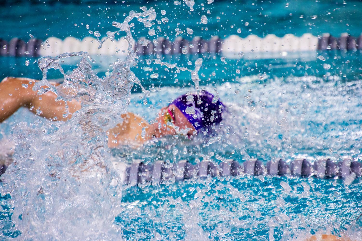 The foreground shows large splashes of water as a person with a purple cap swims in the back.
