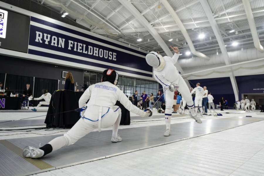 Two people fencing at Ryan Fieldhouse. One is lunging forward, facing away from the camera with the word “Northwestern” written on their back. The other is leaning forward, facing the camera with a white mask over their face. Over the people fencing is a sign with the words “Ryan Fieldhouse” written.