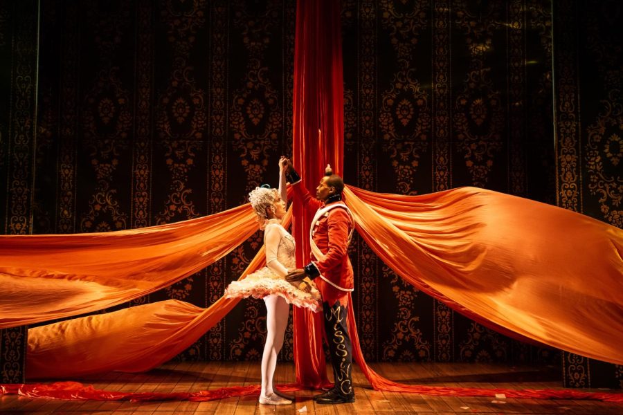 A white woman dressed in a white tutu and tights with a white wig dances with a Black man dressed in a red soldier costume. The word “missing” is written in gold capital lettering along their left leg. Around them hang orange silks and a black and gold patterned background.