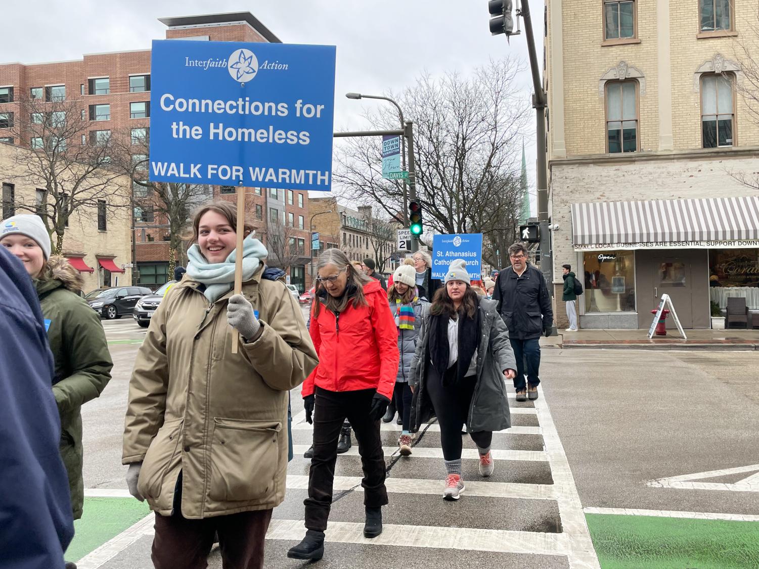 A group of participants walk across the street in downtown Evanston, with one person holding a “Connections for the Homeless” sign. 