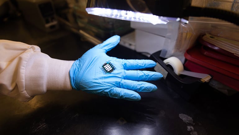A white and gray solar cell in the palm of a hand covered with a blue glove. In the background is a lit desk lamp and a black tape dispenser.
