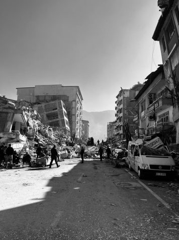 People walk down a road with destroyed buildings on either side. Smashed cars line the street.