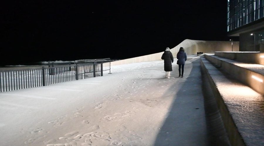Two people walking on a beach covered with snow at night with a glass building next to them.