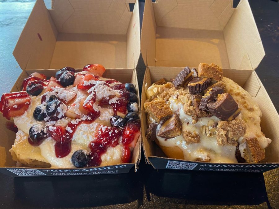 Two cinnamon rolls, one topped with white frosting, brown pie crumble and chocolate pieces, and the other topped with strawberries, blueberries and reddish purple jam.