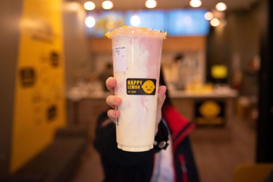A person holding a plastic cup of boba with a black sticker on it that reads “Happy Lemon.”