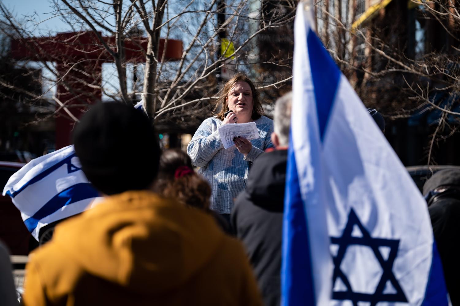 A+woman+reads+from+a+paper+in+front+of+a+crowd+and+an+Israeli+flag.