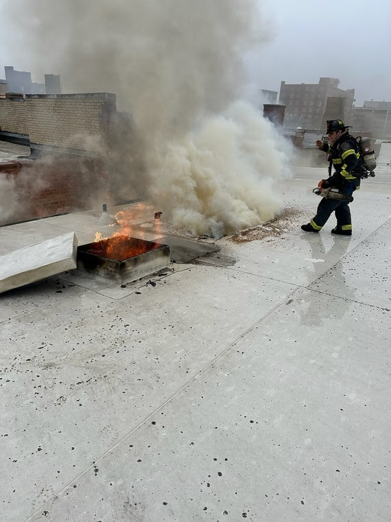 A firefighter facing smoke extinguishes a flame on a rooftop.