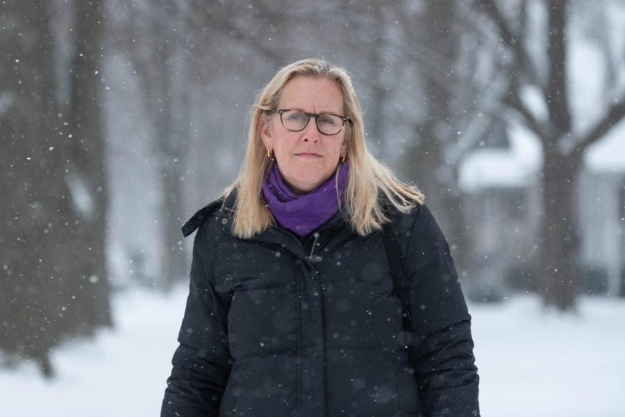 A woman wearing glasses, a black coat and purple scarf looks at the camera while standing outside in the snow.
