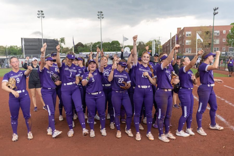 A group of softball players in purple jerseys and purple pants celebrate after a win.
